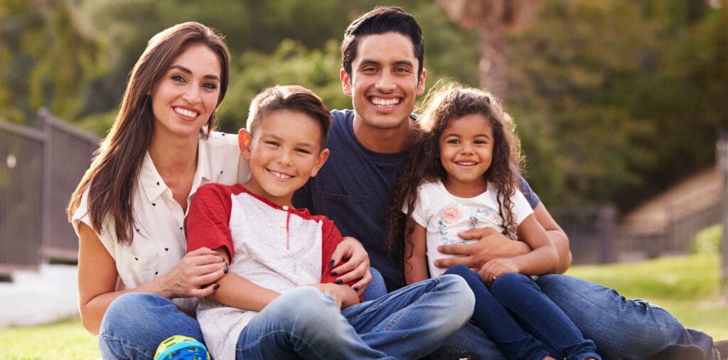 A happy family of four, including two adults and two children, smiling at the camera outdoors. The family consists of a man, a woman, a boy, and a girl.