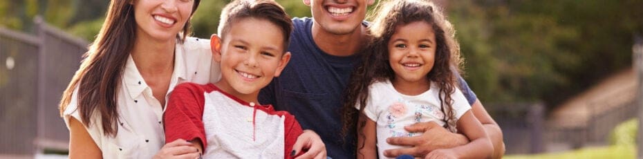 A happy family of four, including two adults and two children, smiling at the camera outdoors. The family consists of a man, a woman, a boy, and a girl.
