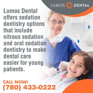 A smiling female dentist or dental professional in blue scrubs stands behind a smiling young girl in a dental chair.